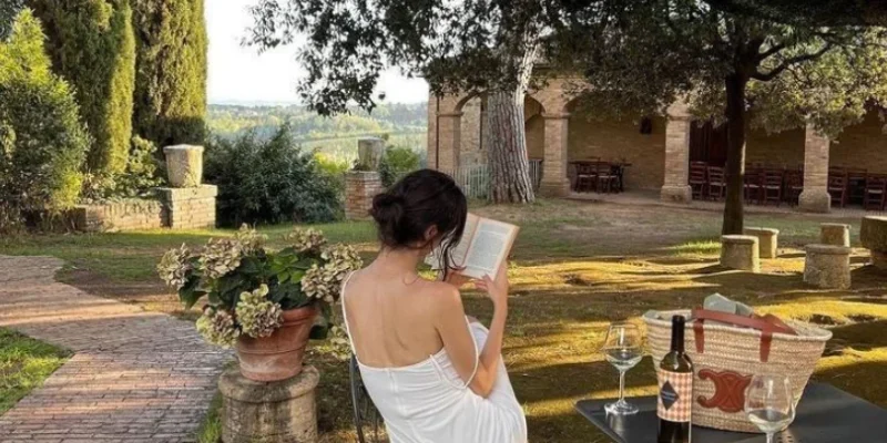 Mujer de espaldas sentada al aire libre leyendo un libro, con una copa de vino sobre la mesa y paisaje campestre al fondo.