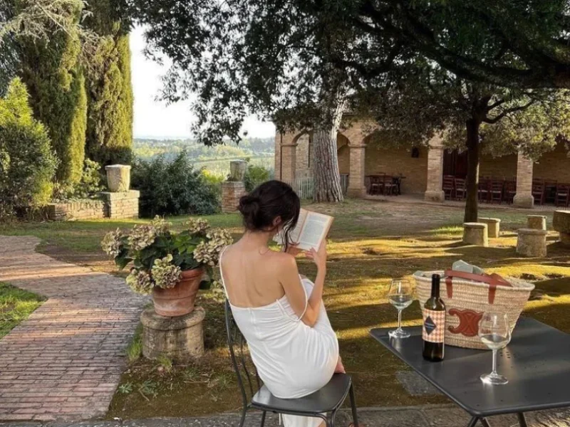 Mujer de espaldas sentada al aire libre leyendo un libro, con una copa de vino sobre la mesa y paisaje campestre al fondo.