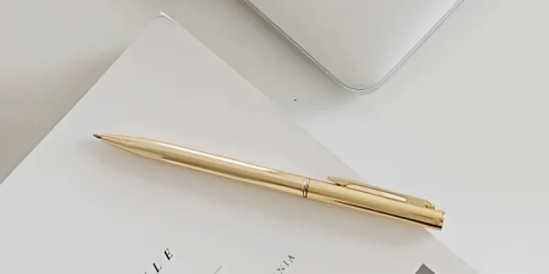 Bolígrafo dorado sobre una revista blanca, junto a una computadora portátil plateada.