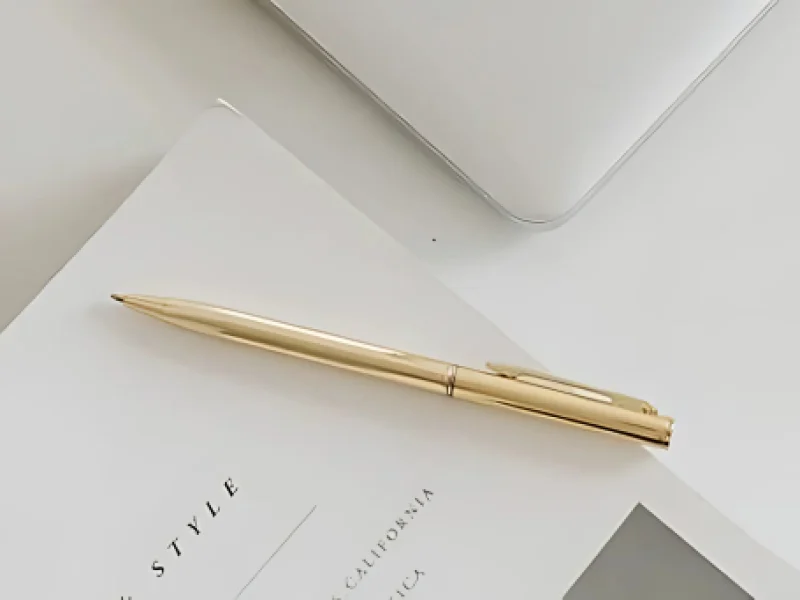 Bolígrafo dorado sobre una revista blanca, junto a una computadora portátil plateada.